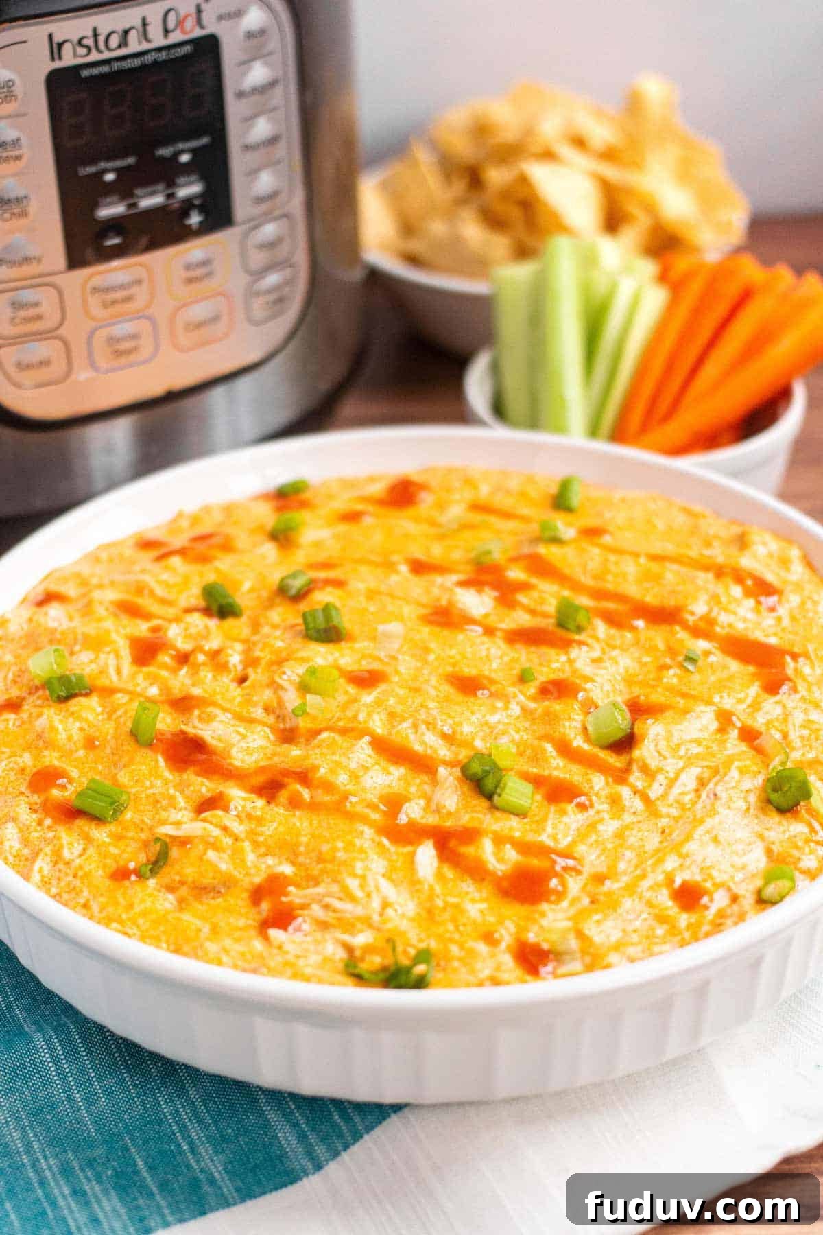 Buffalo chicken dip in a large baking dish garnished with green onions and hot sauce; chips and veggies in background.