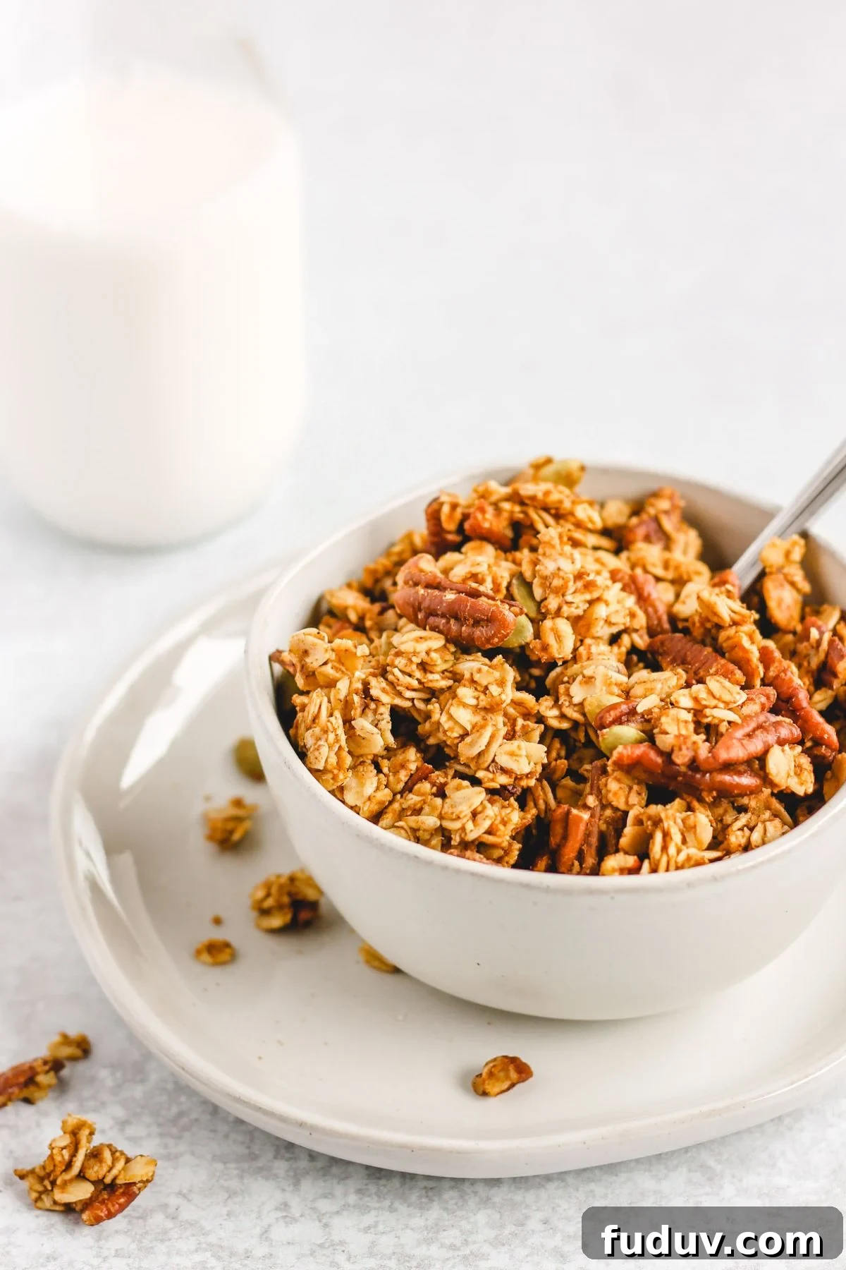 Pumpkin granola in a bowl on a small plate with a glass jar of milk behind bowl.