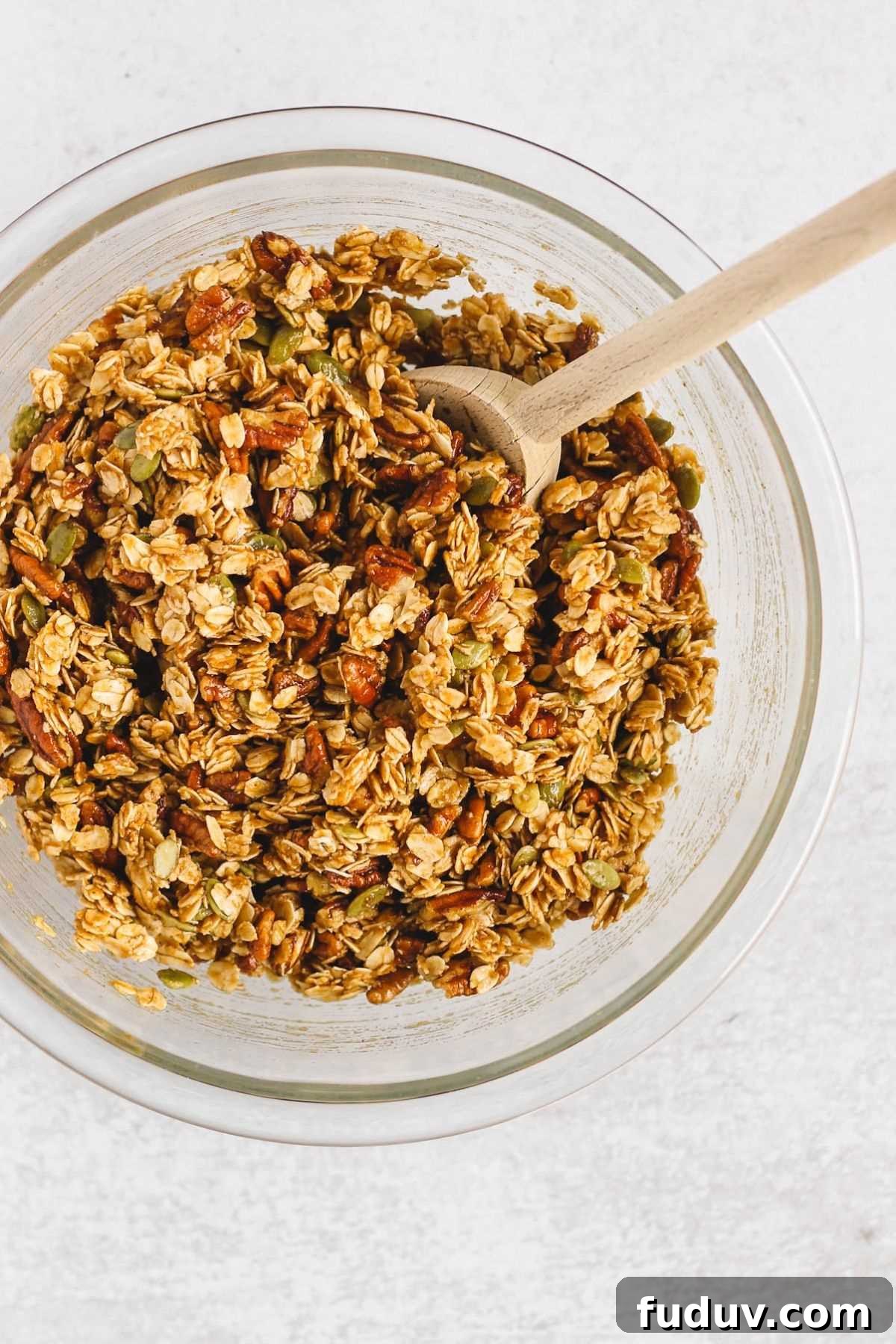 Pumpkin granola ingredients mixed together in a glass mixing bowl with a wooden spoon.