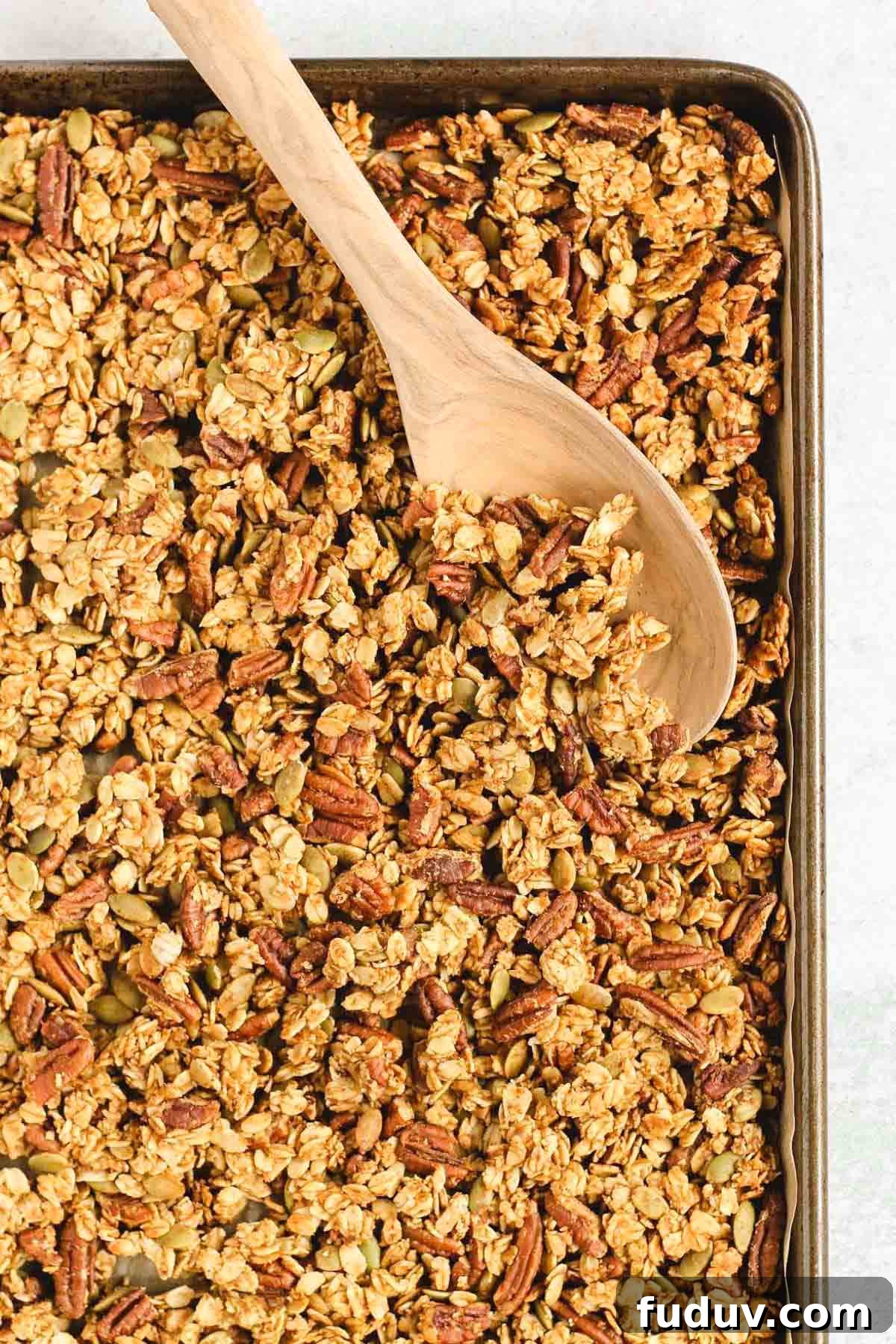 Pumpkin granola on a large baking sheet with a wooden spoon scooping up some granola.