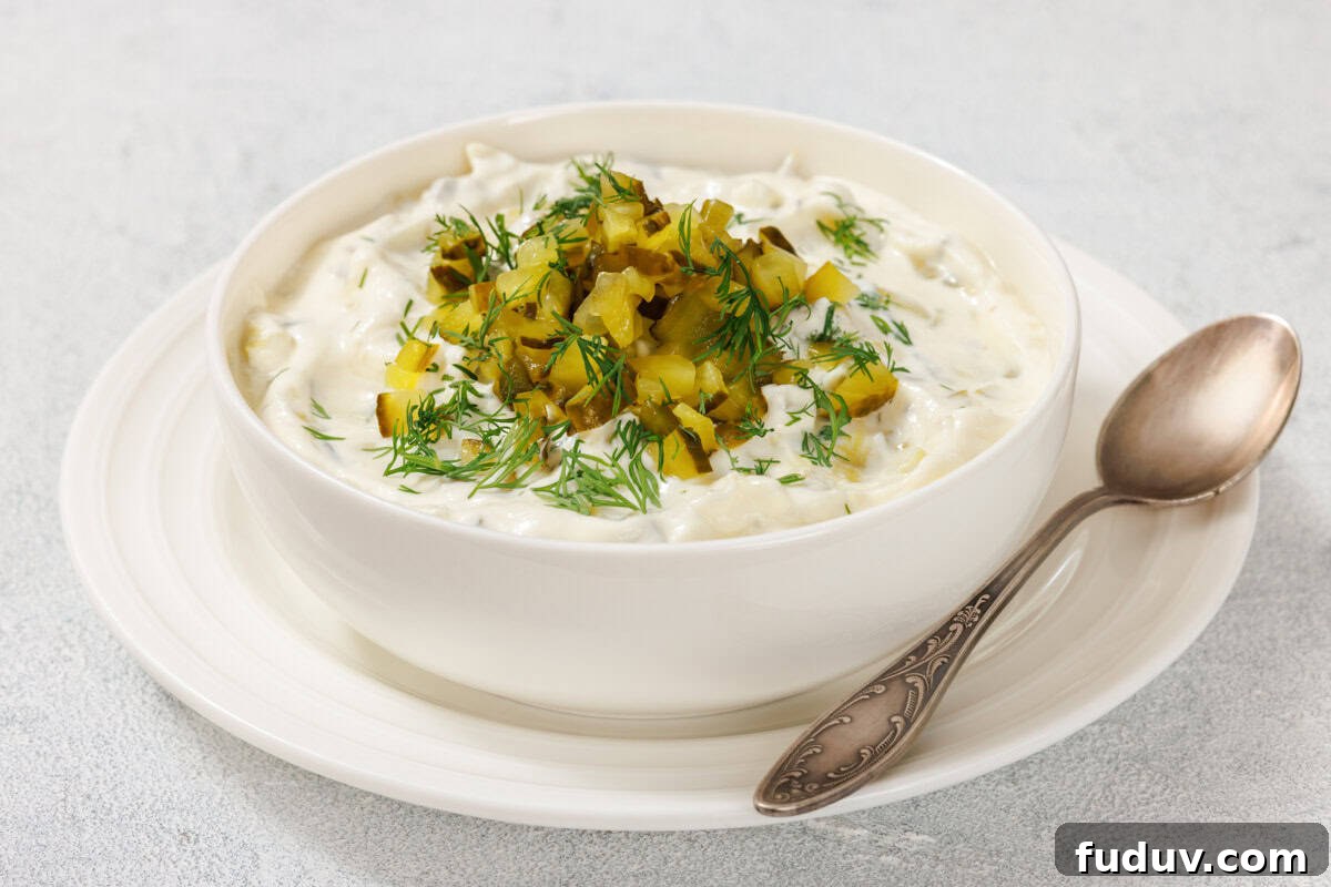 A bowl of Dill Pickle Dip with chopped pickles and dill on top, placed on a white plate. A spoon rests beside the bowl.