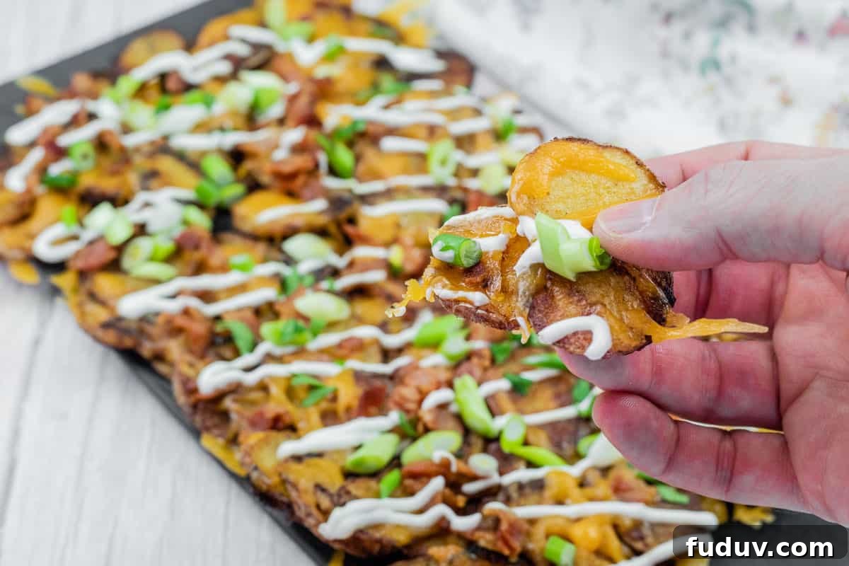 Hand holding a loaded Irish nachos with cheese, bacon, sour cream, and green onions, over a platter of similarly topped slices on a table.