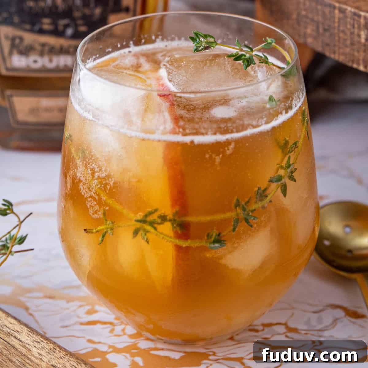 A cocktail in a round glass with ice cubes, garnished with a sprig of thyme and an apple slice. A golden hue liquid fills the glass.