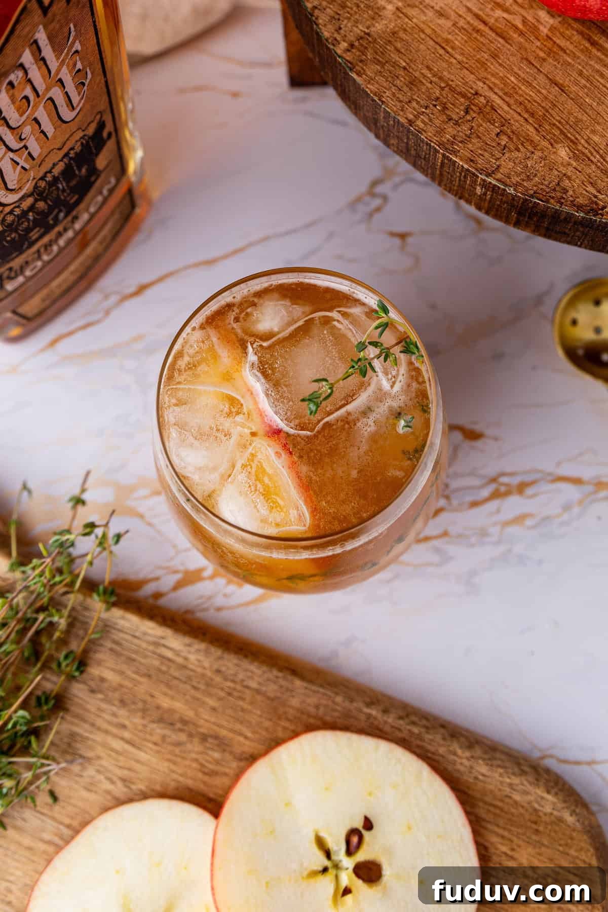 A glass of iced apple cocktail garnished with a sprig of thyme on a marble surface, next to apple slices and fresh thyme.