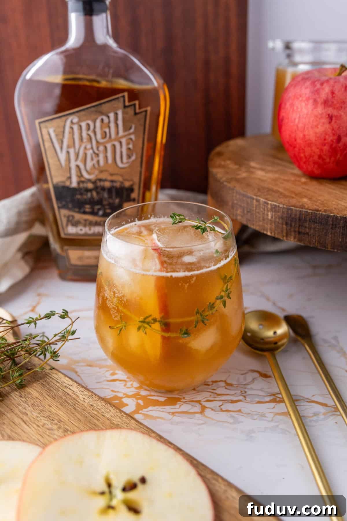 A glass of iced cocktail garnished with thyme and apple slices, placed on a marble surface. A bottle of bourbon, an apple, and gold spoons are nearby.