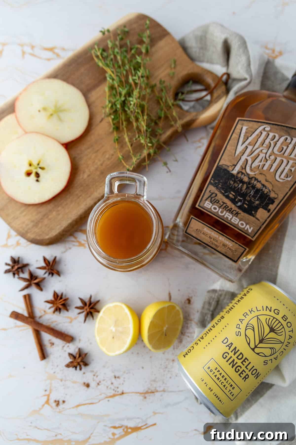 A bottle of bourbon, a mug with a brown liquid, sliced apples, thyme, lemon slices, star anise, cinnamon sticks, and a can of dandelion ginger drink on a marble surface and wooden board.