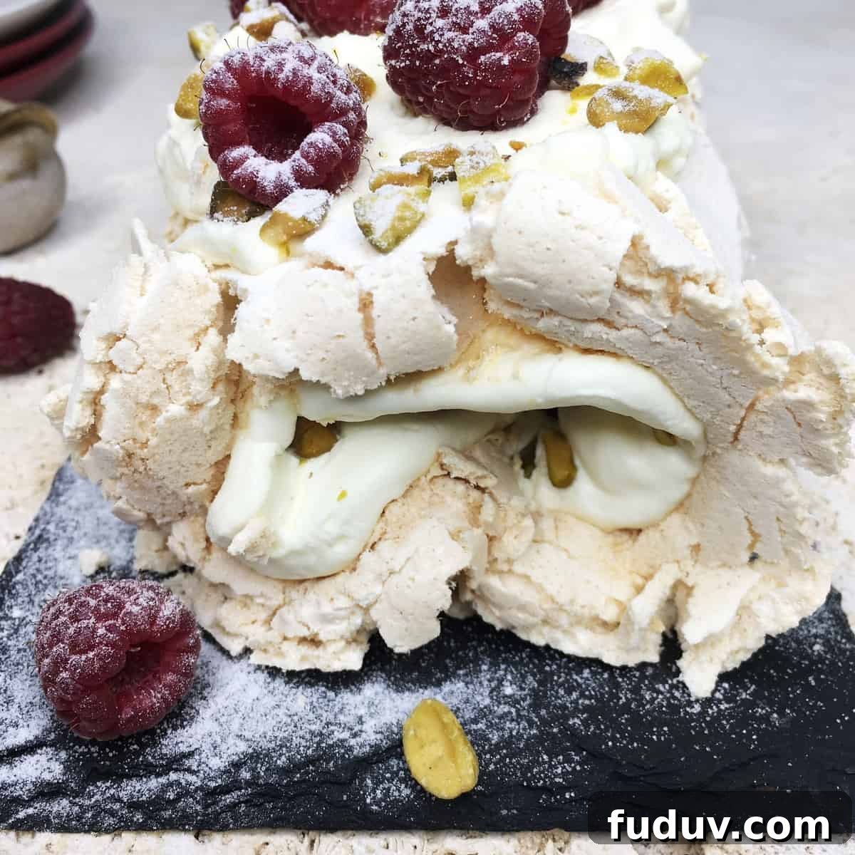 Meringue roulade topped with raspberries and pistachios on a black serving board.