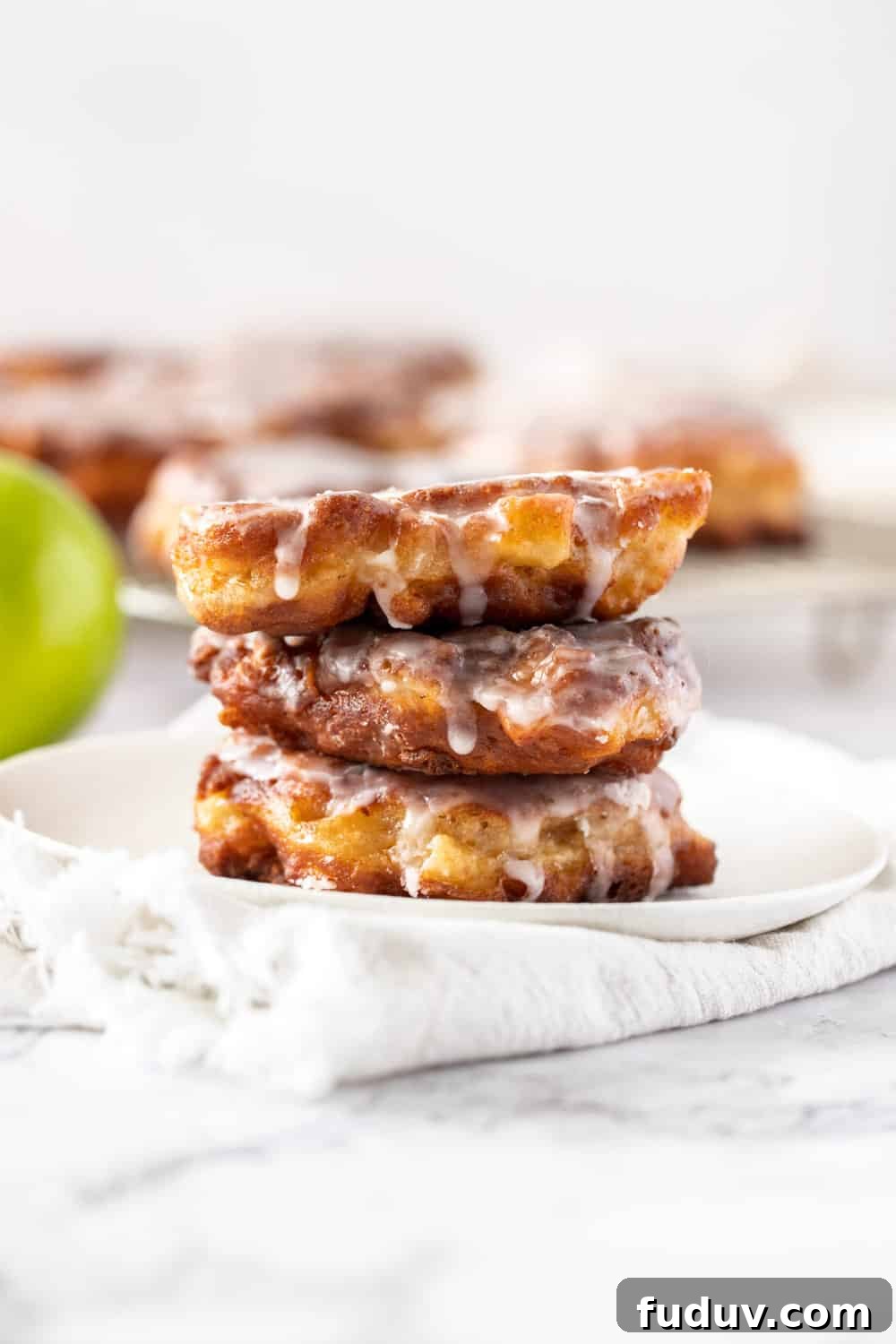 A stack of 3 apple fritters on a white plate with more in the background