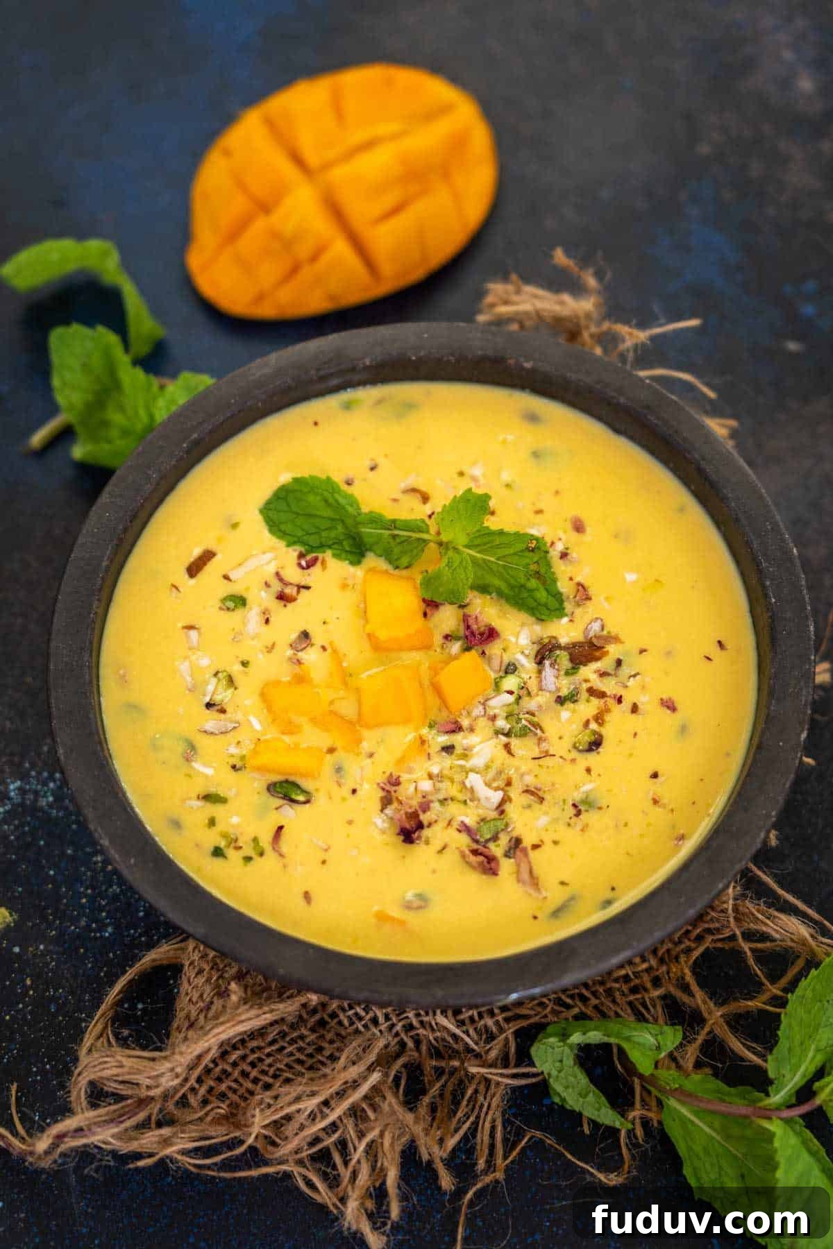 Creamy Mango Basundi Recipe: Authentic Indian Dessert Guide 2 A black bowl filled with creamy Mango Basundi, is garnished with chopped mango pieces, mint leaves, and nuts on a burlap cloth. A sliced mango and fresh mint leaves rest in the background on a dark surface.