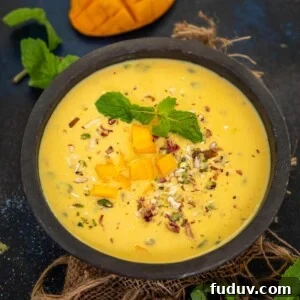 Creamy Mango Basundi Recipe: Authentic Indian Dessert Guide 12 A black bowl filled with creamy Mango Basundi, is garnished with chopped mango pieces, mint leaves, and nuts on a burlap cloth. A sliced mango and fresh mint leaves rest in the background on a dark surface.