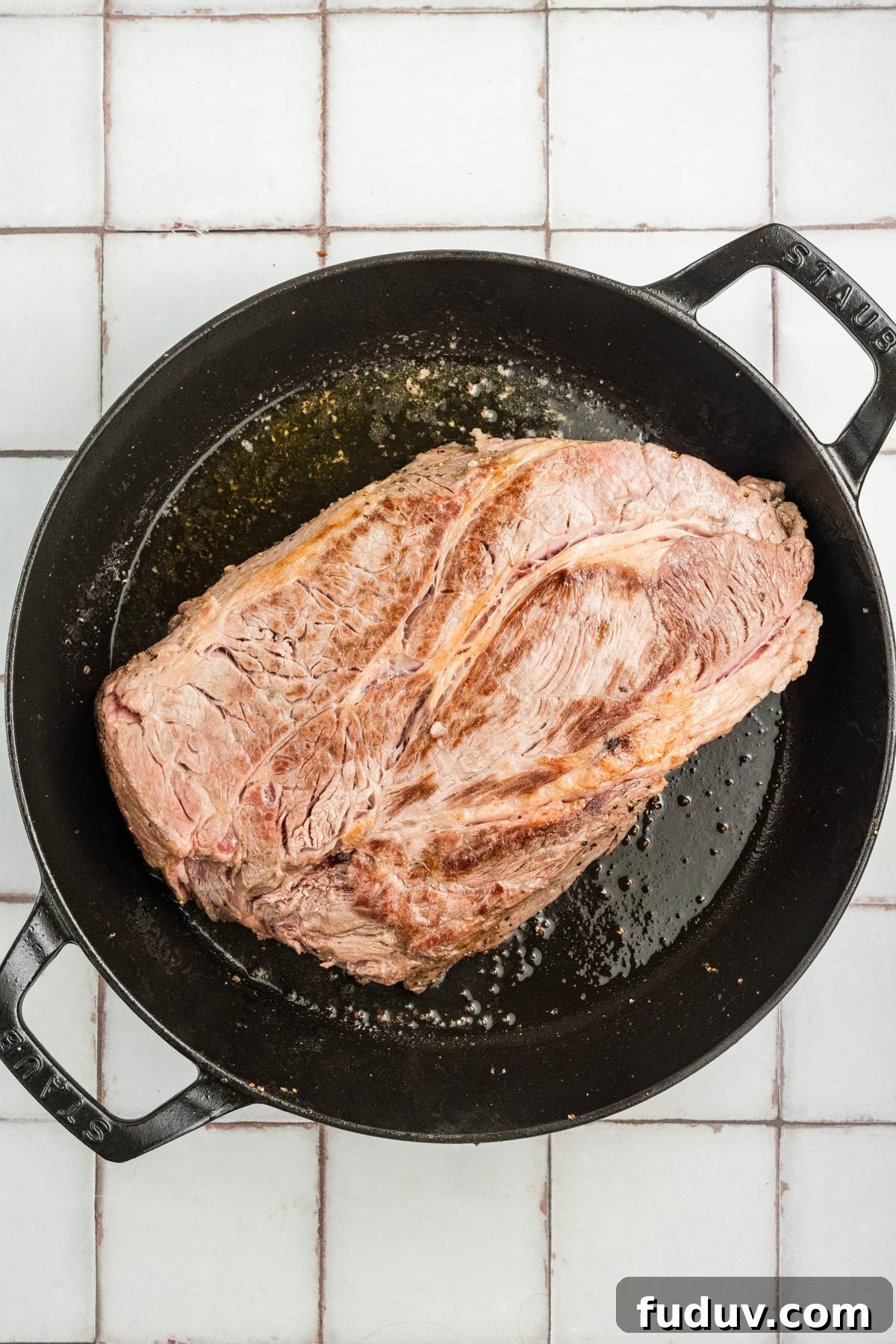 A large piece of seared beef in a black cast iron pot on a white tiled surface.