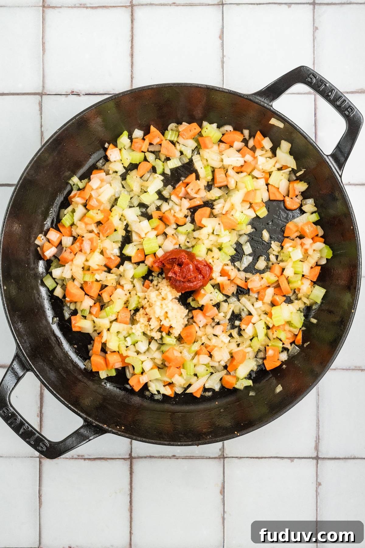 Diced carrots, celery, onion, garlic, and tomato paste in a black skillet on a tiled surface.