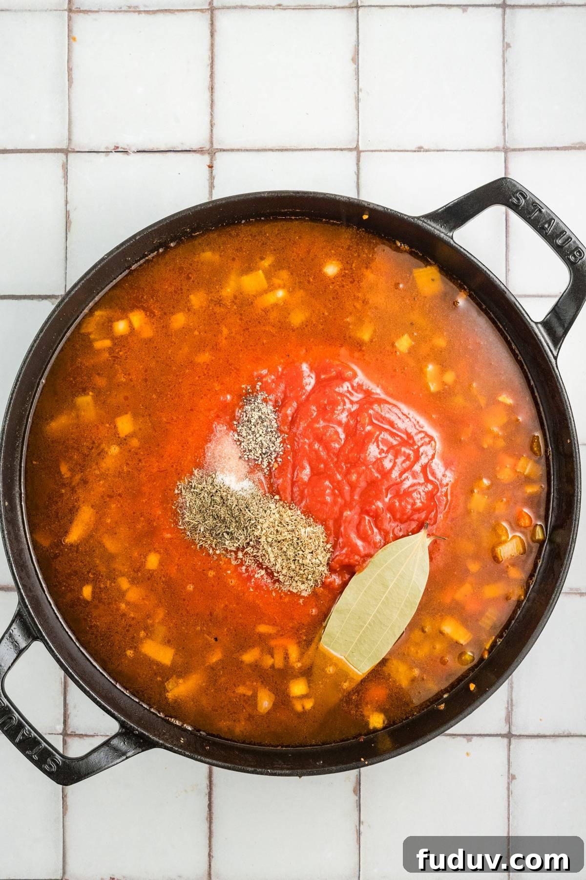 Black pot of soup with diced vegetables, tomatoes, herbs, and a bay leaf on a light tiled surface.