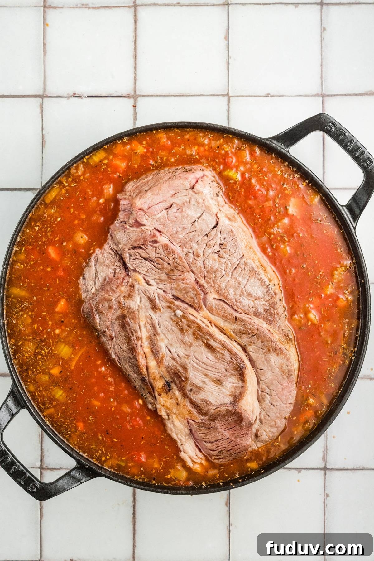 A large cut of beef in a pot of tomato sauce with vegetables on a tiled surface.