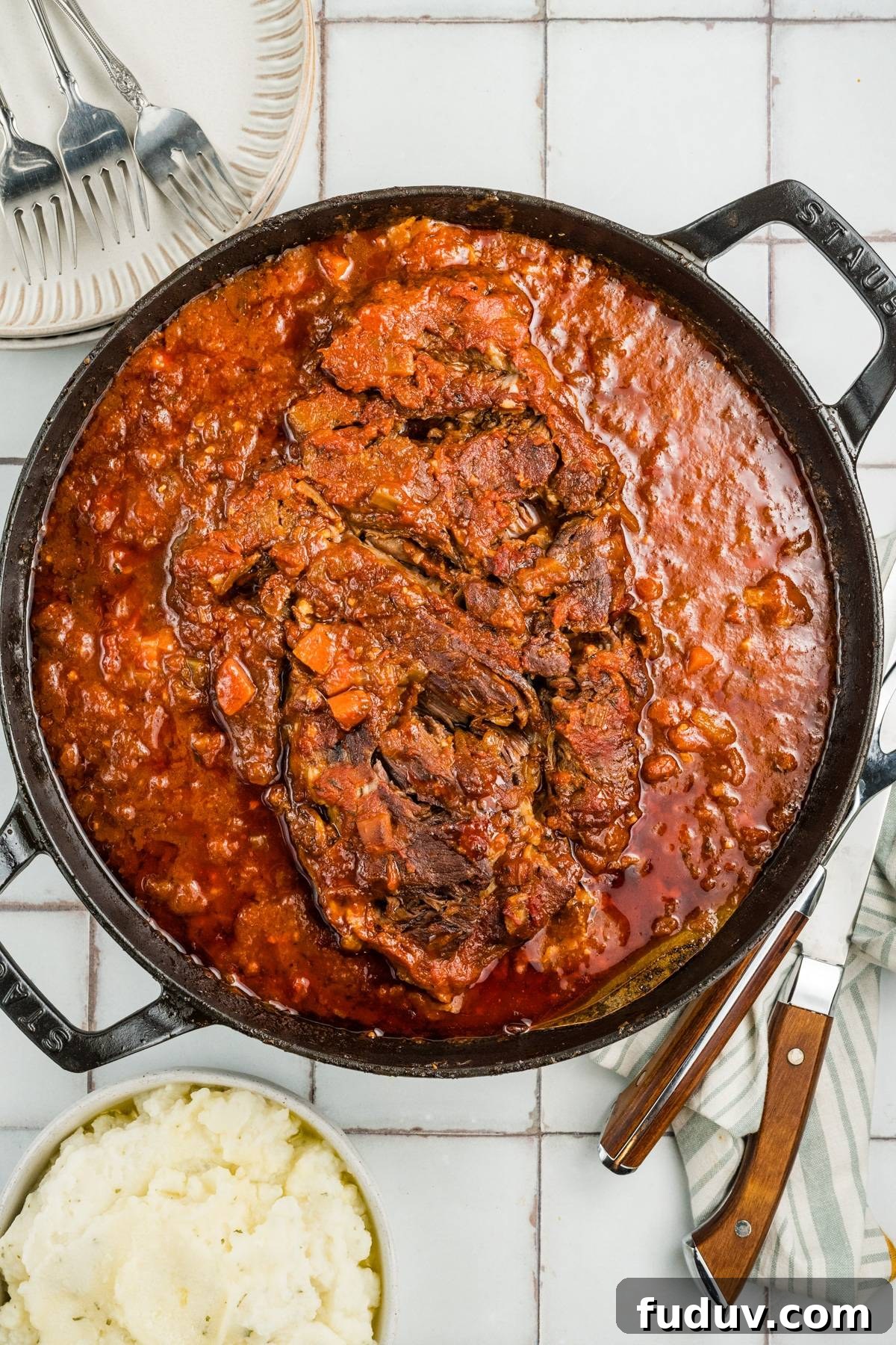 Large pot of braised beef in tomato sauce, accompanied by mashed potatoes, plates, and cutlery on the side.