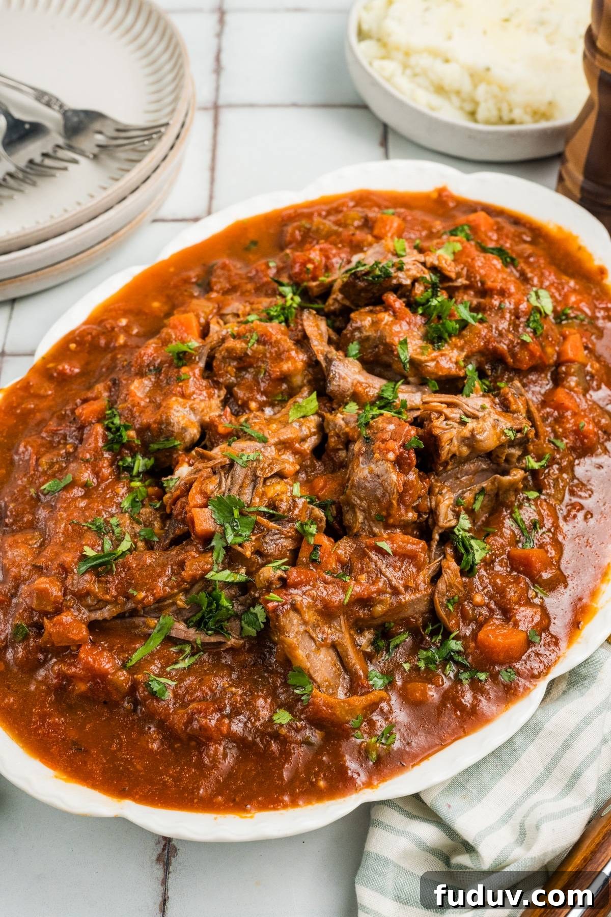 A platter of shredded beef in tomato sauce, garnished with chopped herbs, next to mashed potatoes.