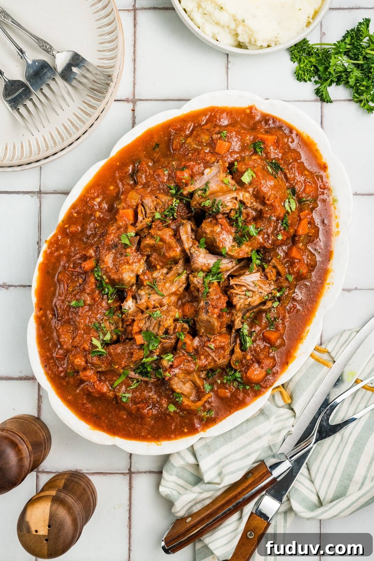 A white plate with beef stew in tomato sauce, garnished with herbs, on a tiled table with utensils nearby.
