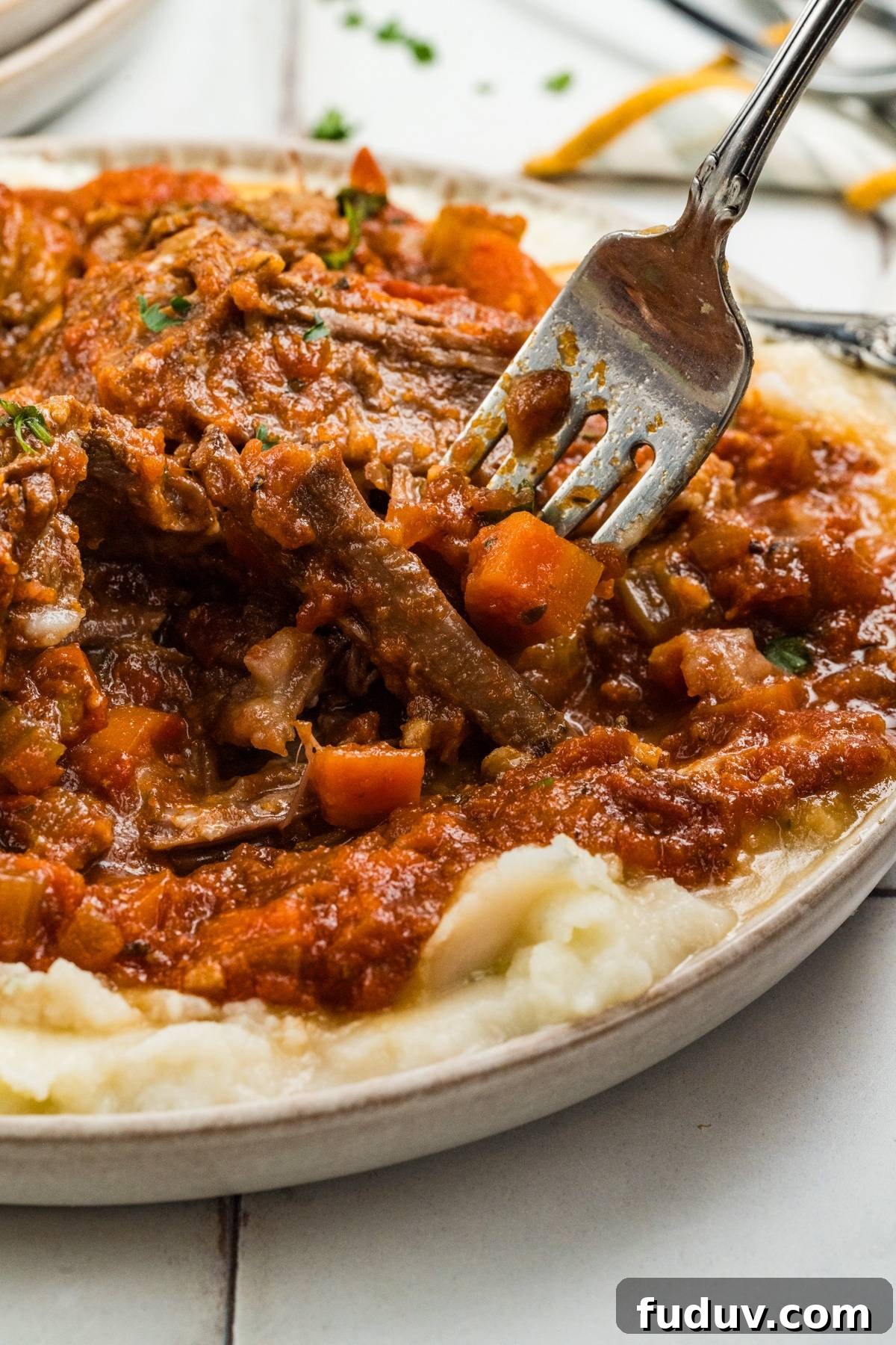 A fork lifts braised beef with vegetables and tomato sauce over creamy mashed potatoes.