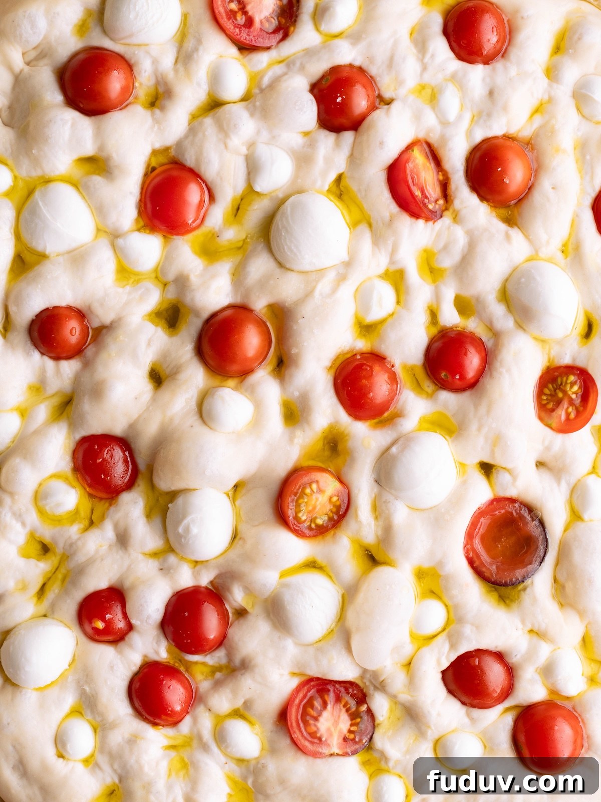 Bubbly focaccia bread dough dimpled with cherry tomatoes, basil flower oil, and mozzarella balls