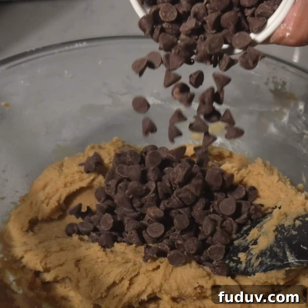 Chewy Chocolate Chip and Gooey Marshmallow Cookies 11 A bowl of cookie dough with chocolate chips being poured in from a white measuring cup, ready for mixing.