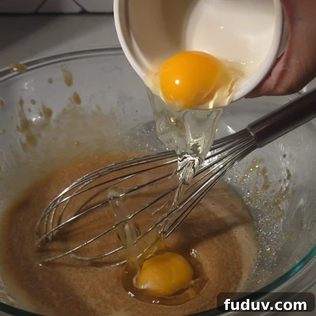 Chewy Chocolate Chip and Gooey Marshmallow Cookies 9 An egg being poured from a white bowl into a glass mixing bowl with a whisk resting inside, showcasing the wet ingredient preparation.