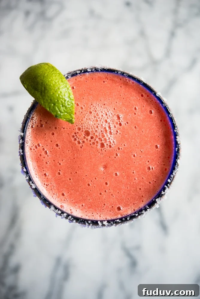 Bright pink frozen watermelon margarita in a blue-rimmed margarita glass with a lime wedge on a marble countertop.