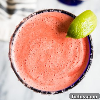 Bright pink frozen watermelon margarita in a blue-rimmed margarita glass with a lime wedge on a marble countertop.