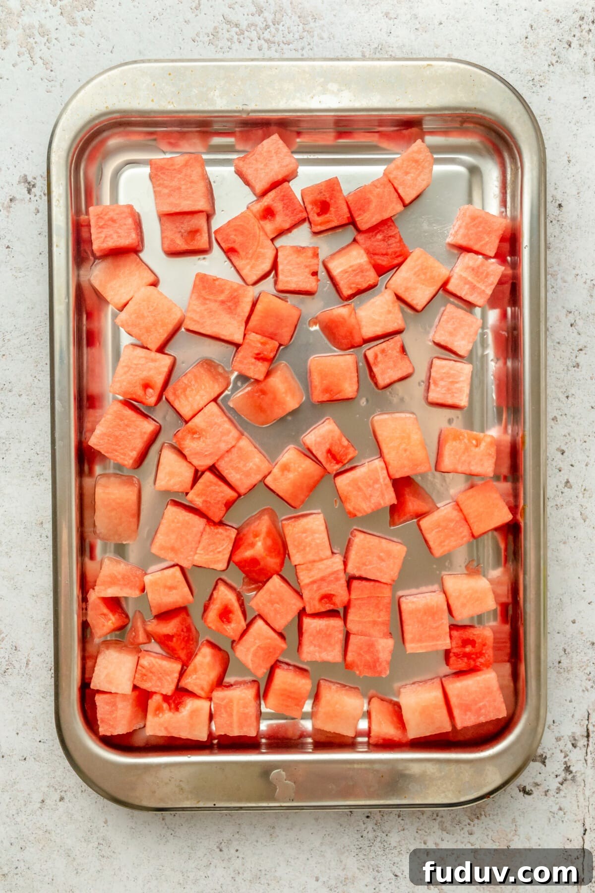 Cubes of fresh watermelon sit on a tray to be frozen on a light grey surface.