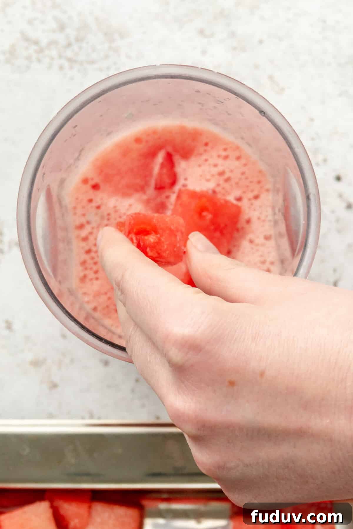 Cubes of frozen margaritas are tossed into a blender beaker for a watermelon margaritas on a light grey surface.
