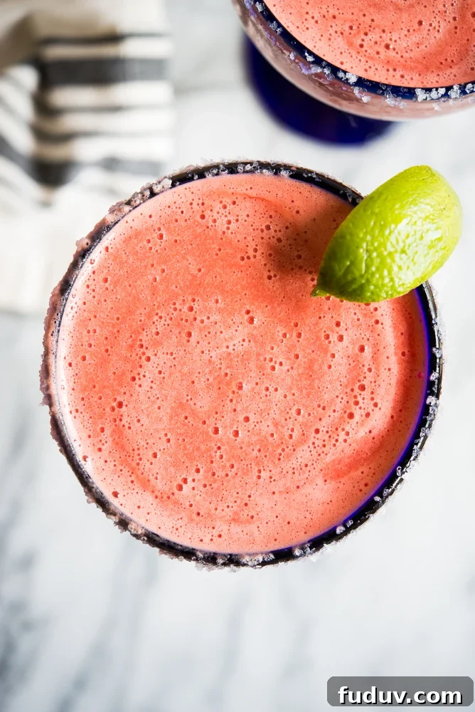 Bright pink frozen watermelon margarita in a blue-rimmed margarita glass with a lime wedge on a marble countertop.