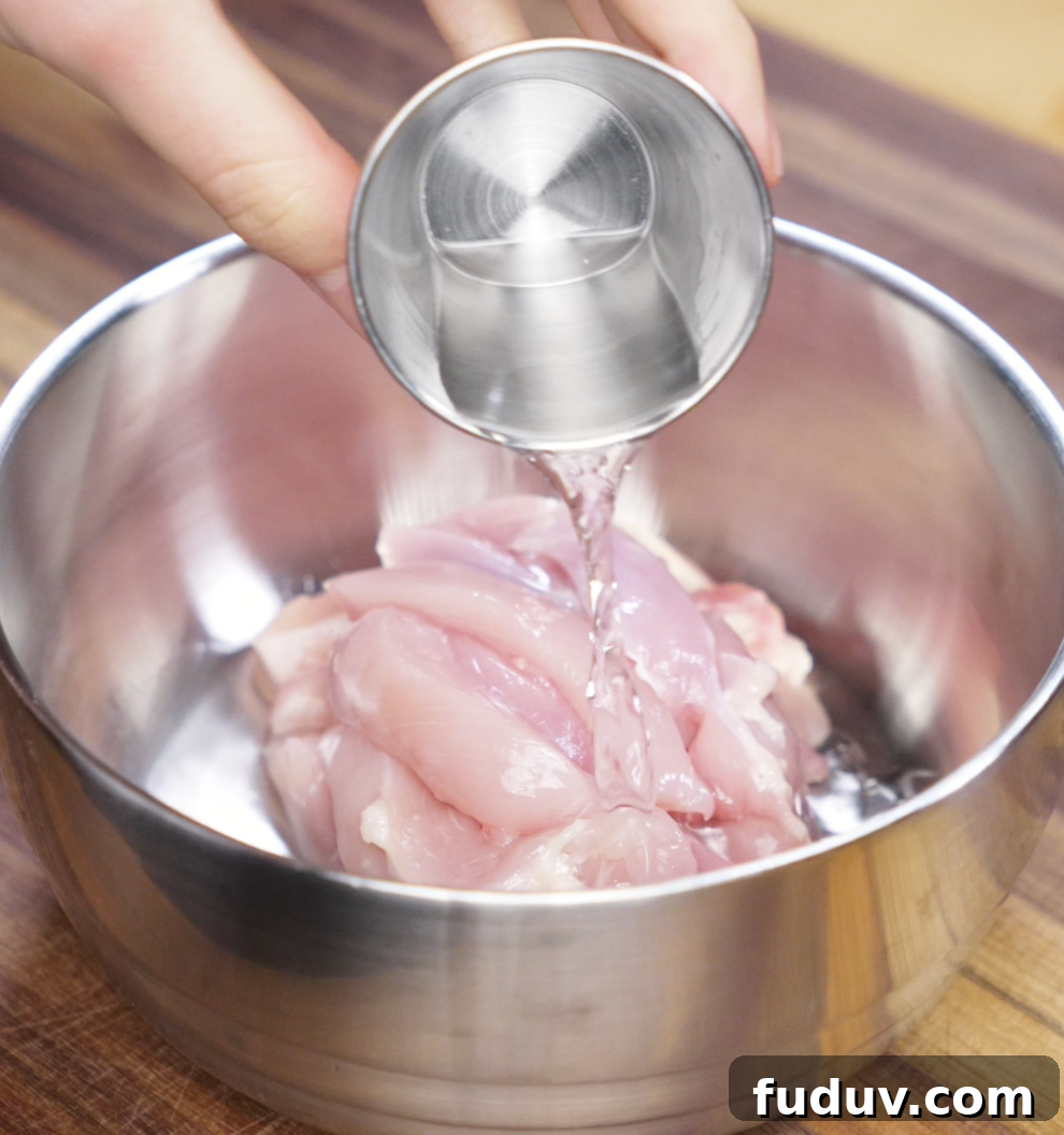 Crispy Chicken Pan-Fried Noodles with Savory Stir-Fry Sauce 6 A hand pours Shaoxing wine over raw sliced chicken thighs in a metal bowl. This step helps tenderize the meat and infuse it with classic Chinese flavor.