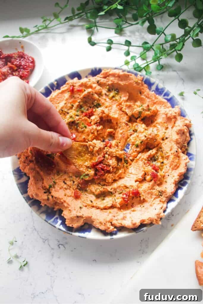 Hand dipping pita chip in sundried tomato feta dip on a small blue and white plate.
