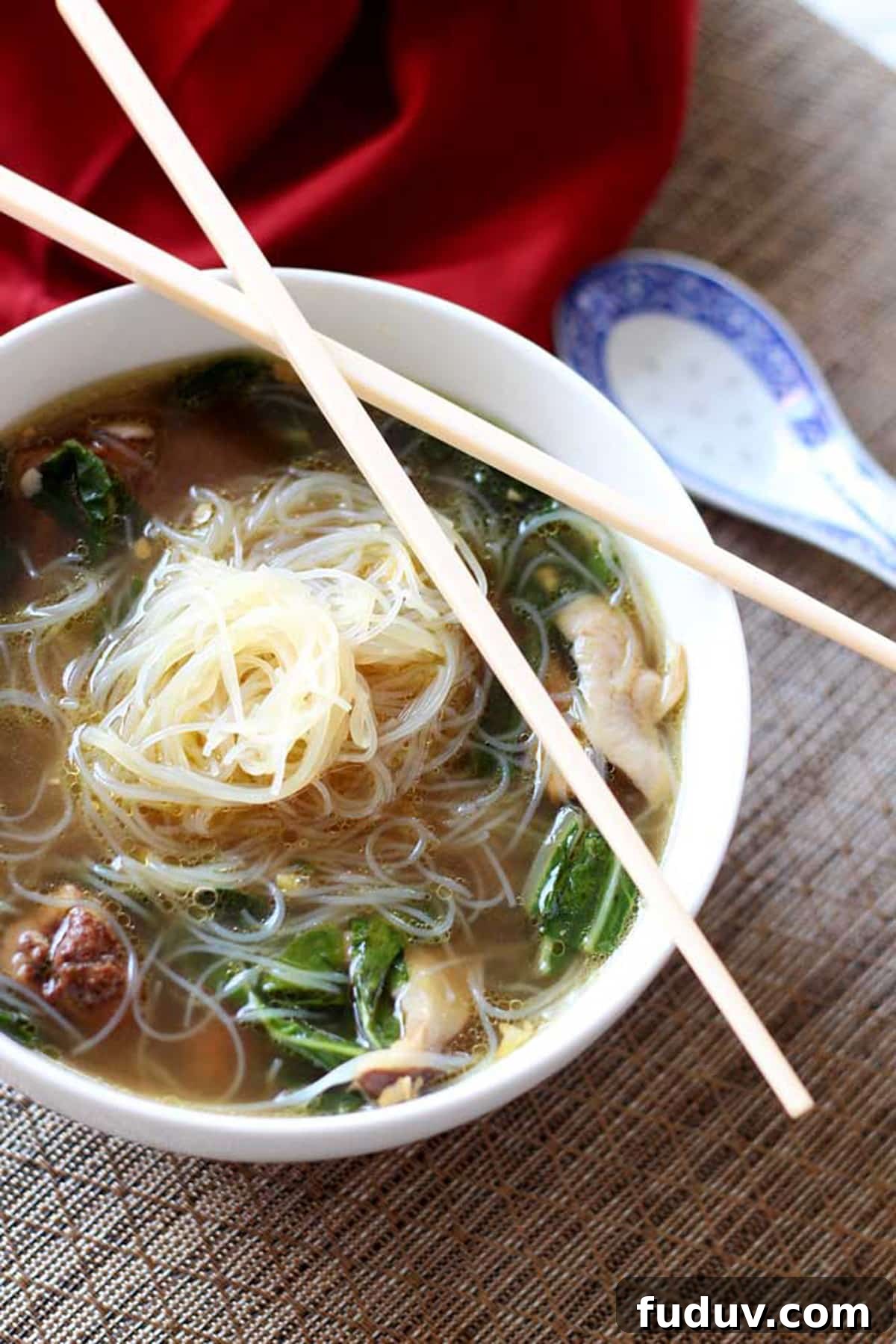 White bowl containing Asian Chicken Noodle Soup with crossed chopsticks sitting on a brown table mat, red napkin on table.