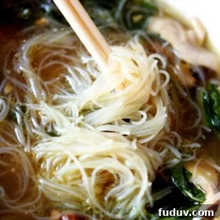 White bowl of Asian Chicken Noodle Soup with Mei Fun Noodles twirled on chopsticks, sitting on a brown table mat.