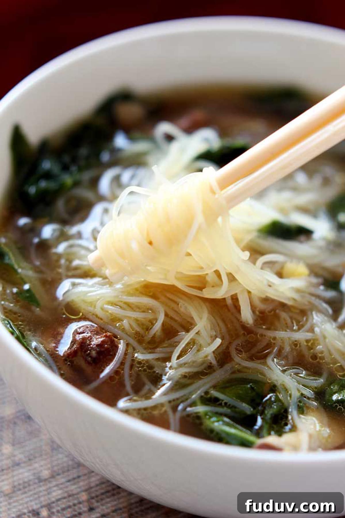 White bowl containing Asian Chicken Noodle soup, white chopsticks twirling Mei Fun noodles.