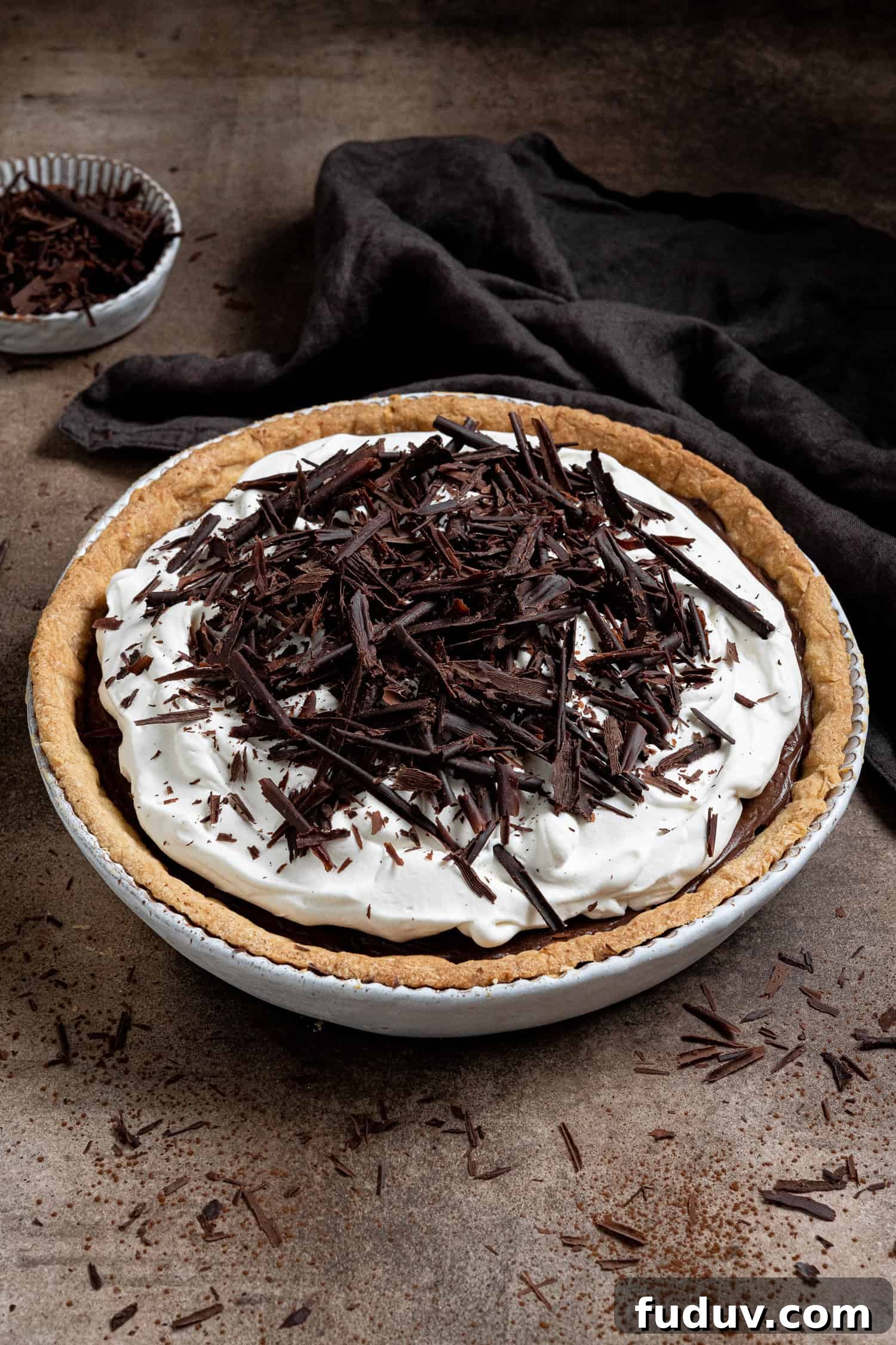 Vegan chocolate cream pie on a brown surface with a bowl of chocolate shavings and a brown cloth.