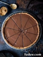 Vegan peanut butter chocolate tart cut into slices on a dark background with chocolate curls, plates, forks, a bowl of peanut butter and a brown cloth.