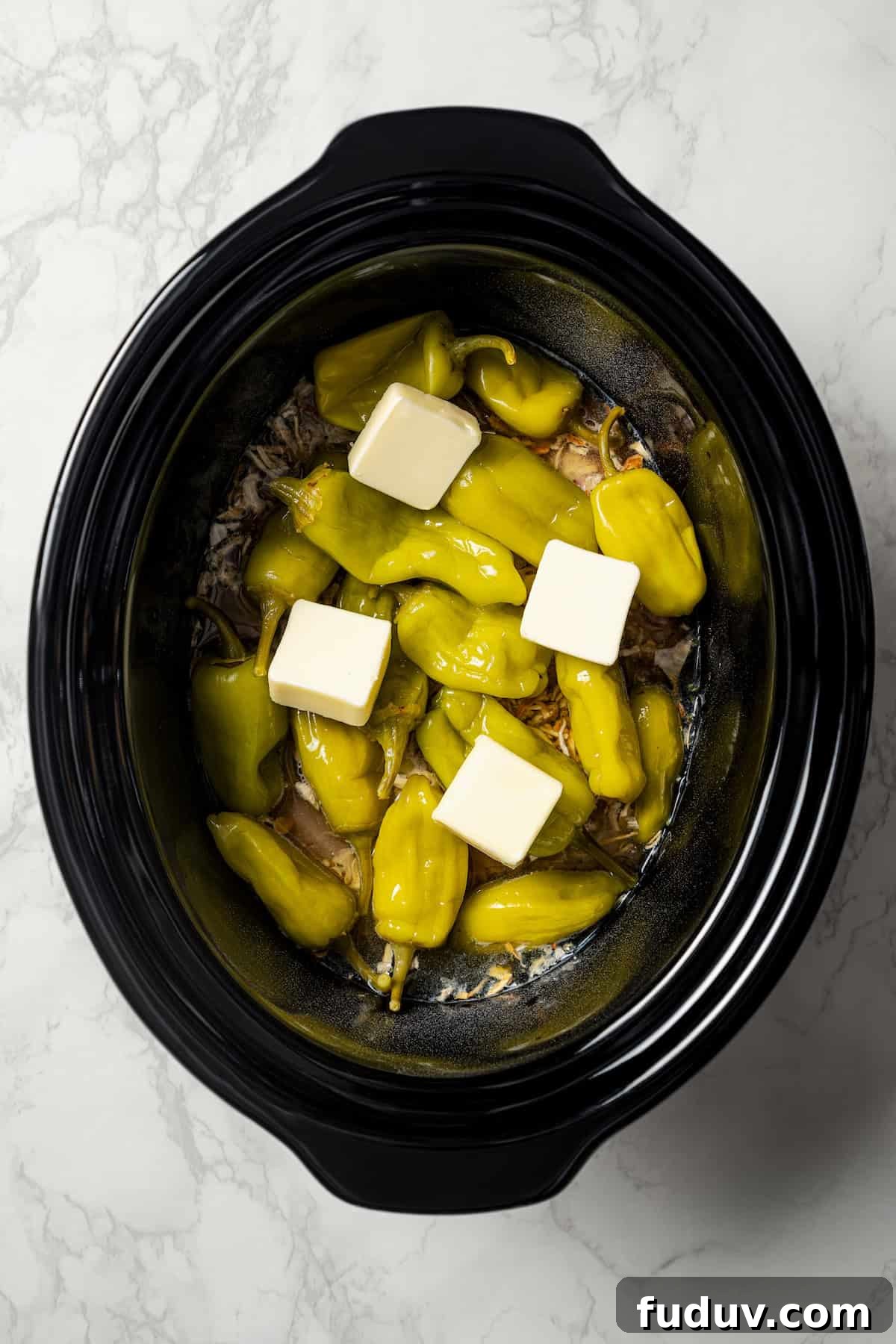 Mississippi chicken is an easy crock pot recipe with juicy chicken shredded in savory pepperoncini ranch sauce. Make it with 5 ingredients! Pats of butter placed over top pepperoncini peppers and chicken inside the crock pot.