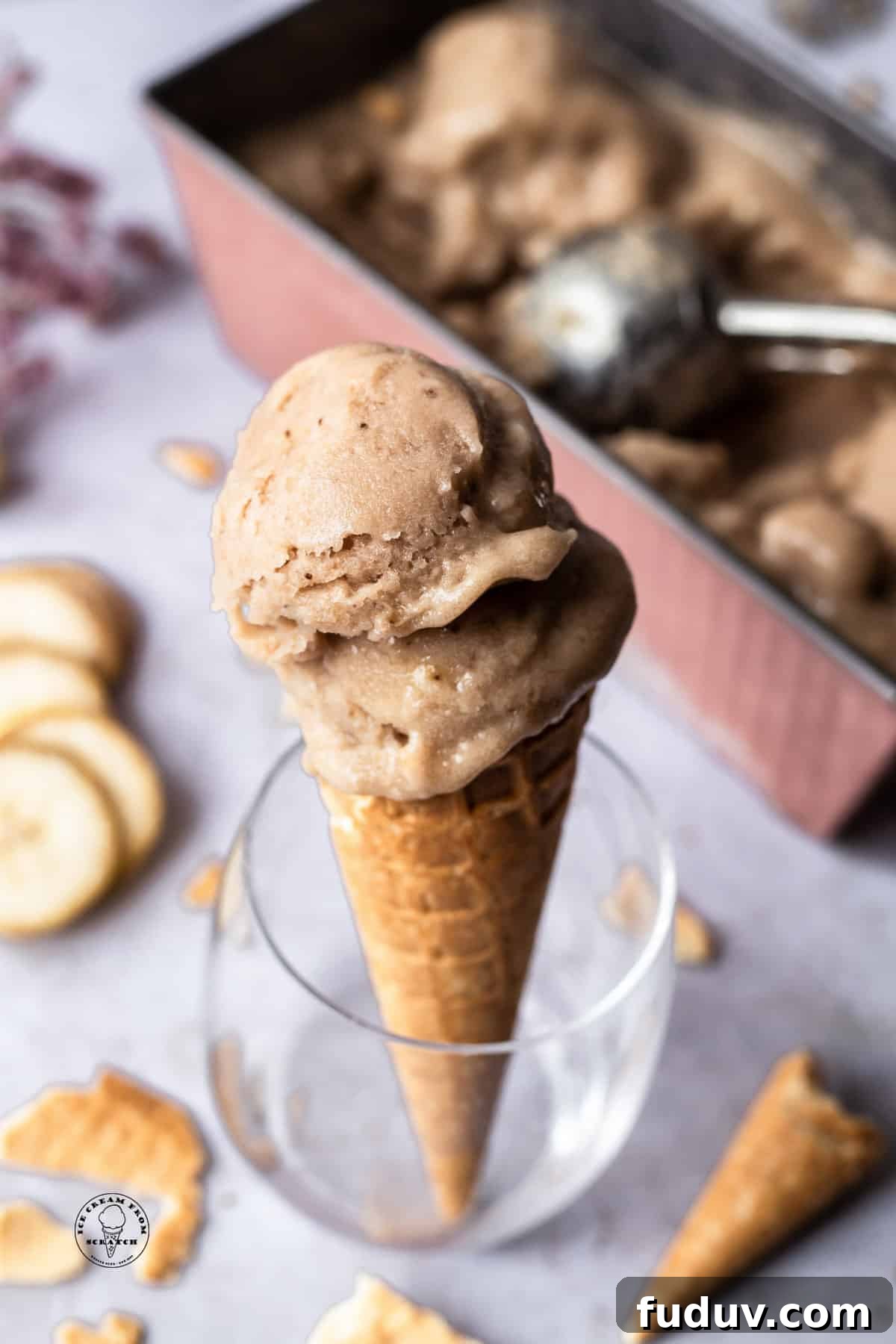 Creamy Banana Ice Cream Recipe: No-Added-Sugar Frozen Treat 2 homemade banana ice cream scooped into a sugar cone and propped up in a glass. in the background is the pan of ice cream, banana slices, and pieces of cones.