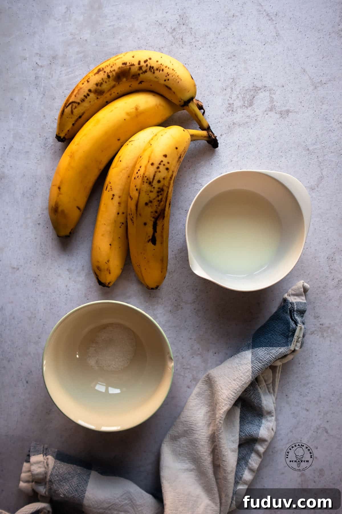 Creamy Banana Ice Cream Recipe: No-Added-Sugar Frozen Treat 3 4 ripe bananas, a small bowl of milk, and a small bowl with a pinch of salt, on a concrete counter. A blue plaid dish towel is there as well.