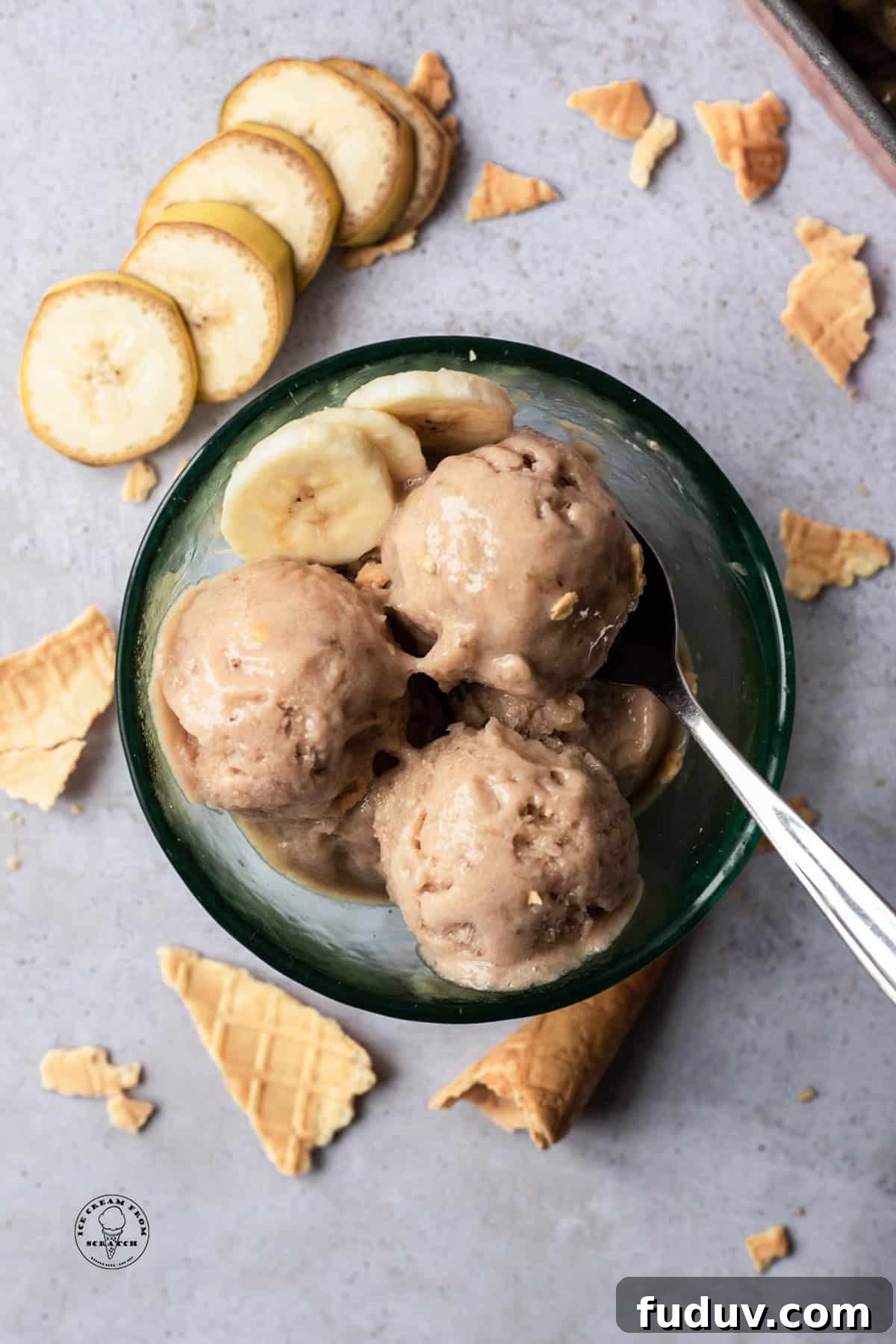 Creamy Banana Ice Cream Recipe: No-Added-Sugar Frozen Treat 6 Scoops of banana ice cream (aka nice cream) in a glass bowl, viewed from overhead. Around the bowl are sliced bananas and pieces of ice cream cones.
