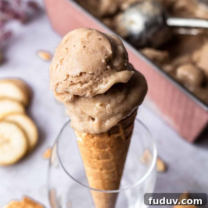 homemade banana ice cream scooped into a sugar cone and propped up in a glass. in the background is the pan of ice cream, banana slices, and pieces of cones.