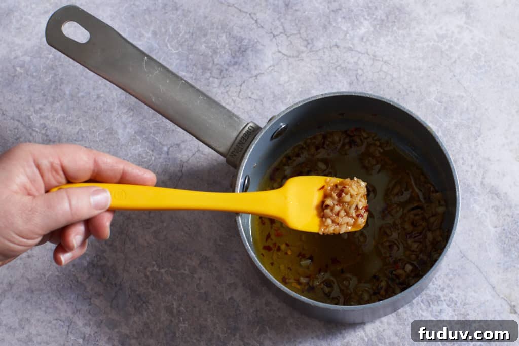 A woman's hand holding a yellow spatula over a small sauce pan with chopped garlic and red pepper.