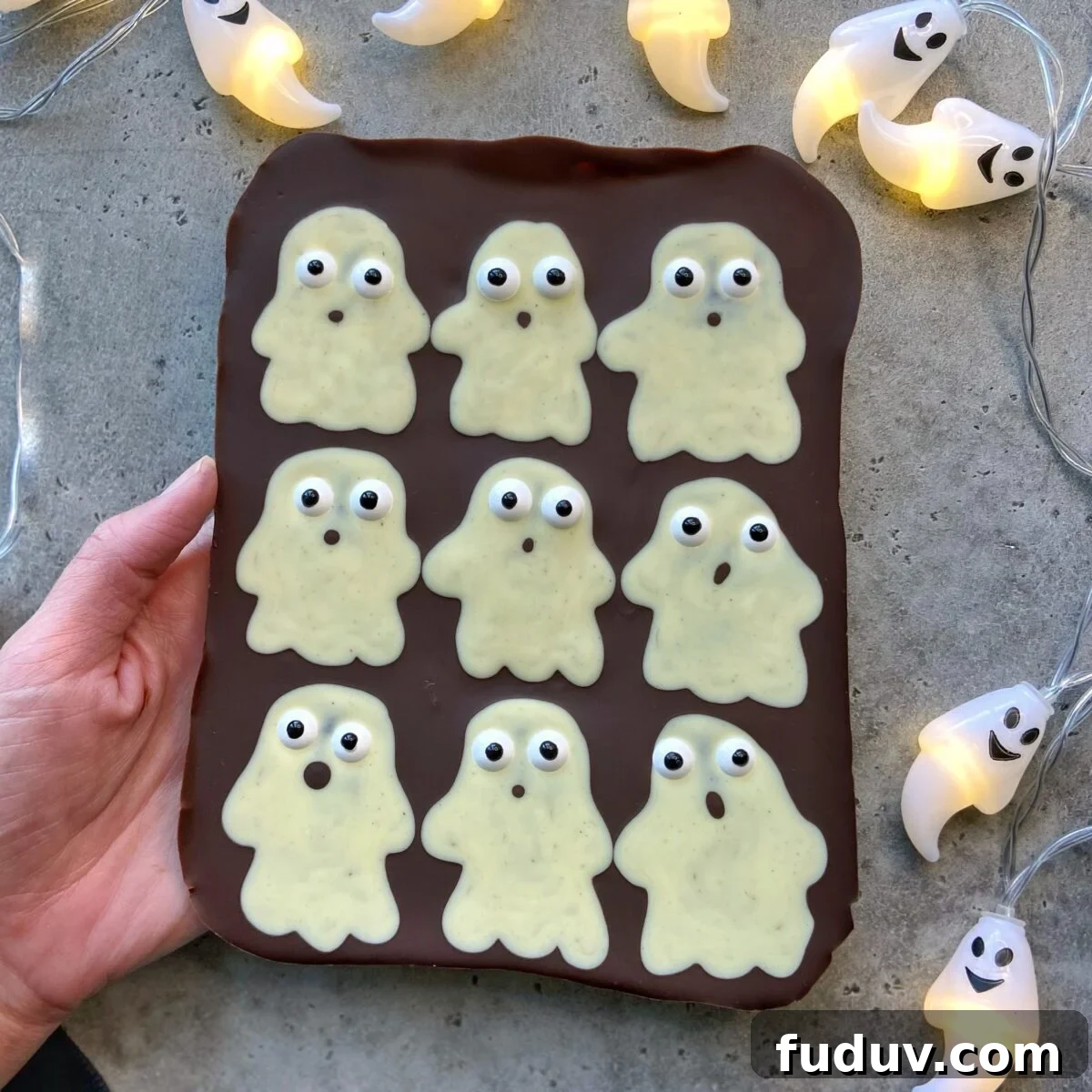 A hand holds a chocolate slab topped with nine white chocolate ghost bites, each with candy eyes, surrounded by ghost-shaped string lights on a gray surface.