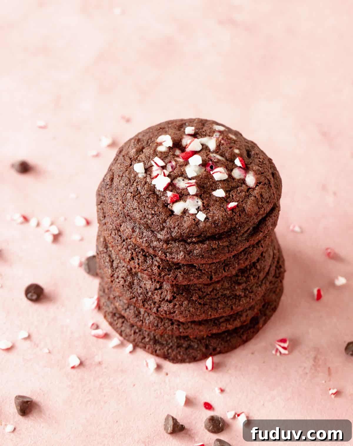 Several chocolate peppermint cookies stacked on pink surface, crushed candy cane and chips around.