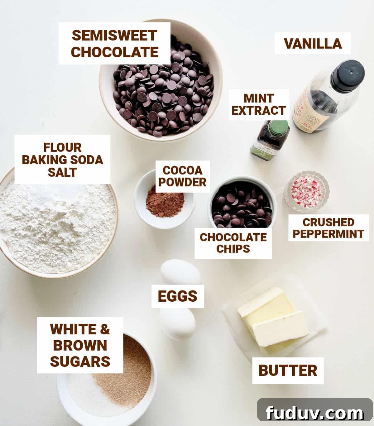 White surface with bowls containing ingredients for peppermint chocolate cookies including butter, flour, sugar, eggs, cocoa, extracts, chips.