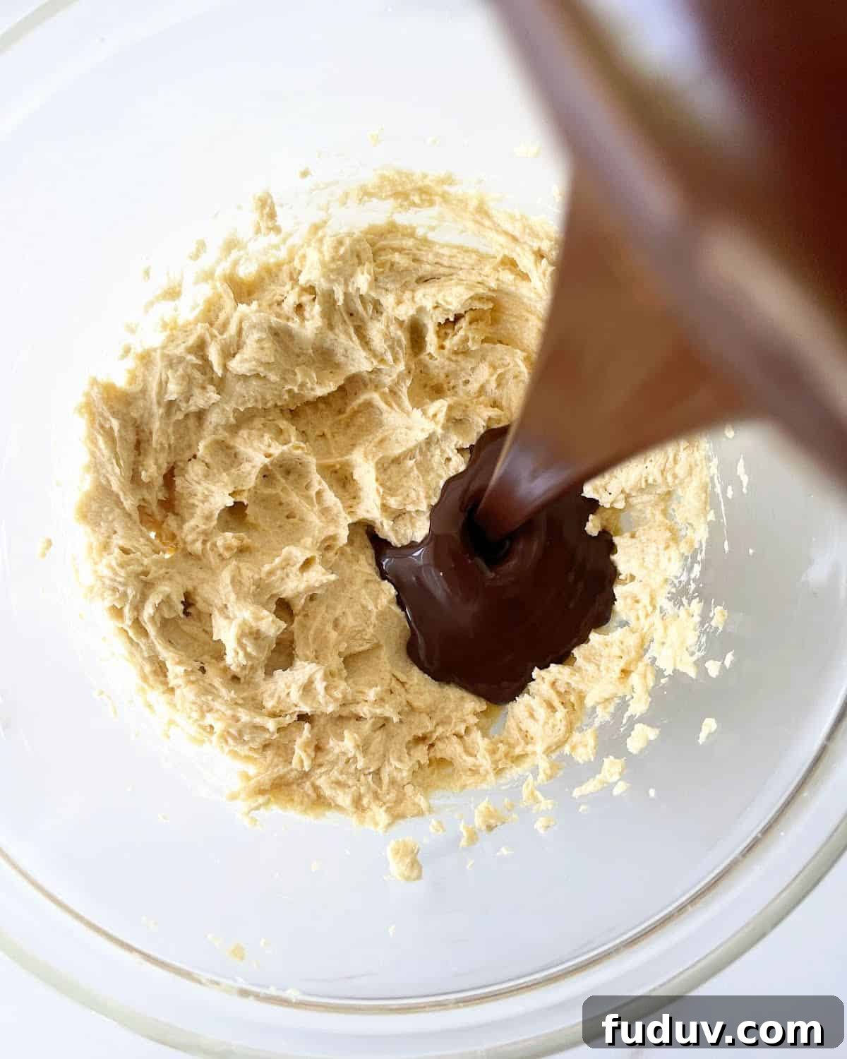 Pouring melted chocolate over butter sugar mixture in glass bowl on a white surface.