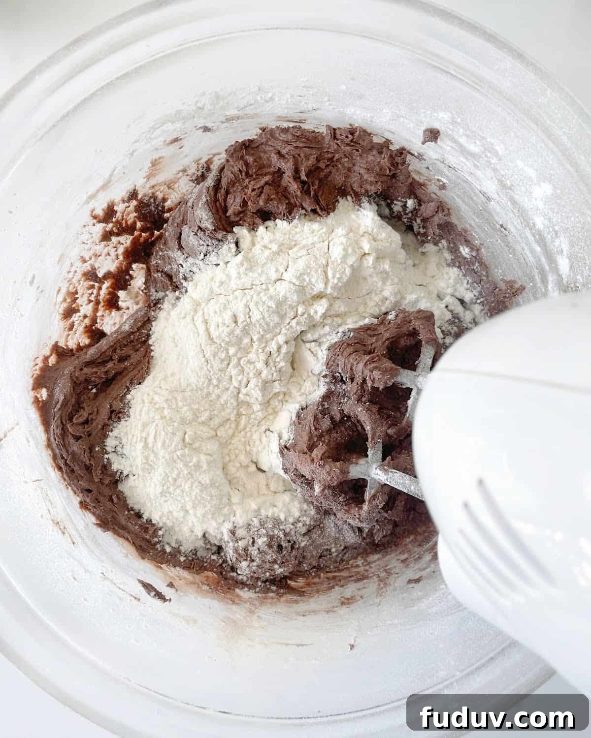 Flour added to chocolate cookie mixture in a glass bowl with an electric mixer. White marble surface.
