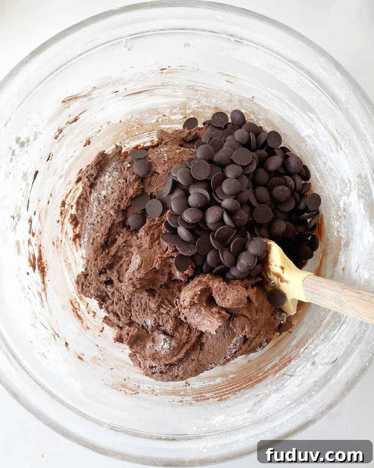 Chocolate chips added to chocolate cookie batter in a glass bowl with a spatula. White marble surface.
