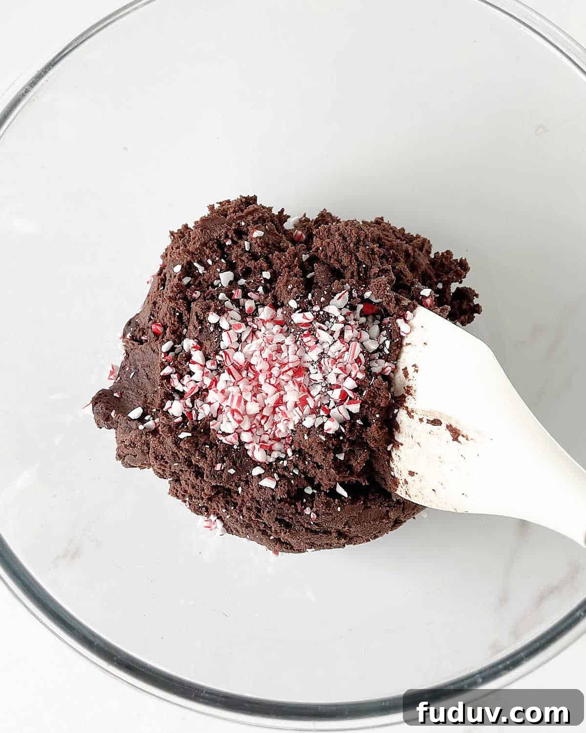Crushed peppermint candy added to chocolate cookie dough on a glass bowl with a white spatula. White marble surface.